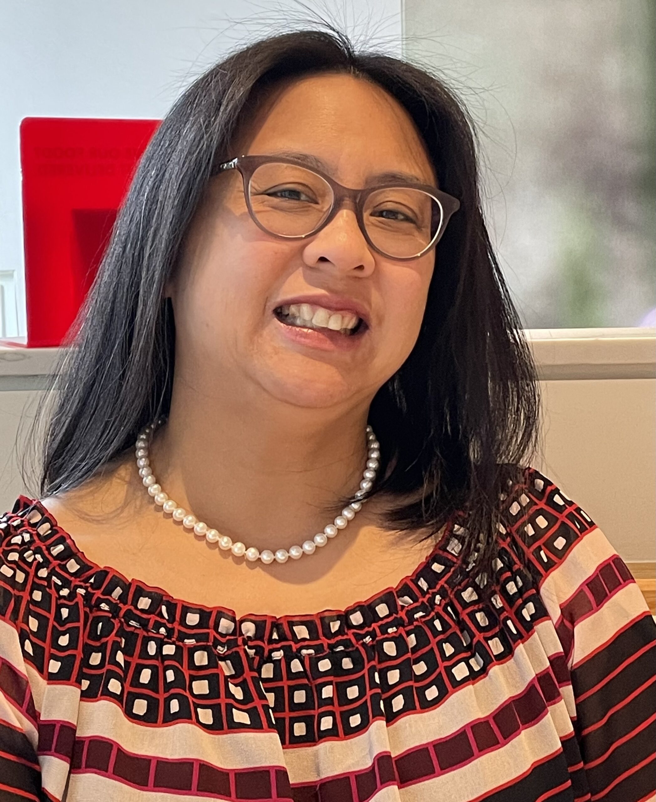 Smiling woman with glasses and long dark hair, wearing a pearl necklace and a red-patterned top with geometric designs. Bright, cheerful ambiance.