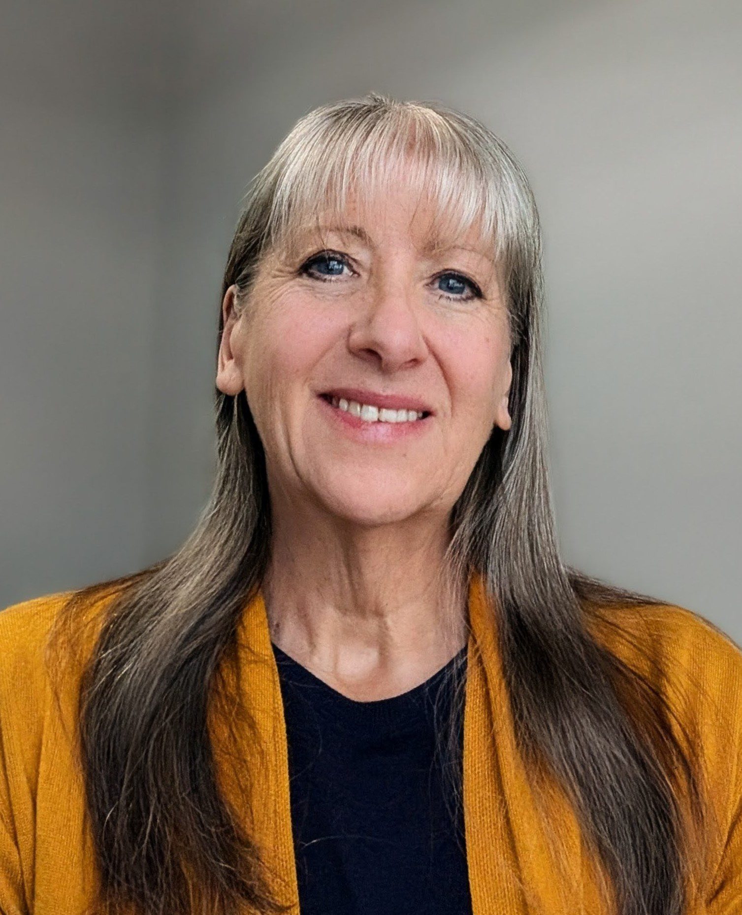 Smiling woman with long gray hair and bangs, wearing a mustard yellow cardigan over a dark top, against a plain gray background.